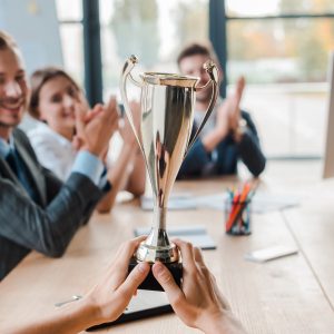 cropped view of businesswoman holding champion cup near coworkers in office